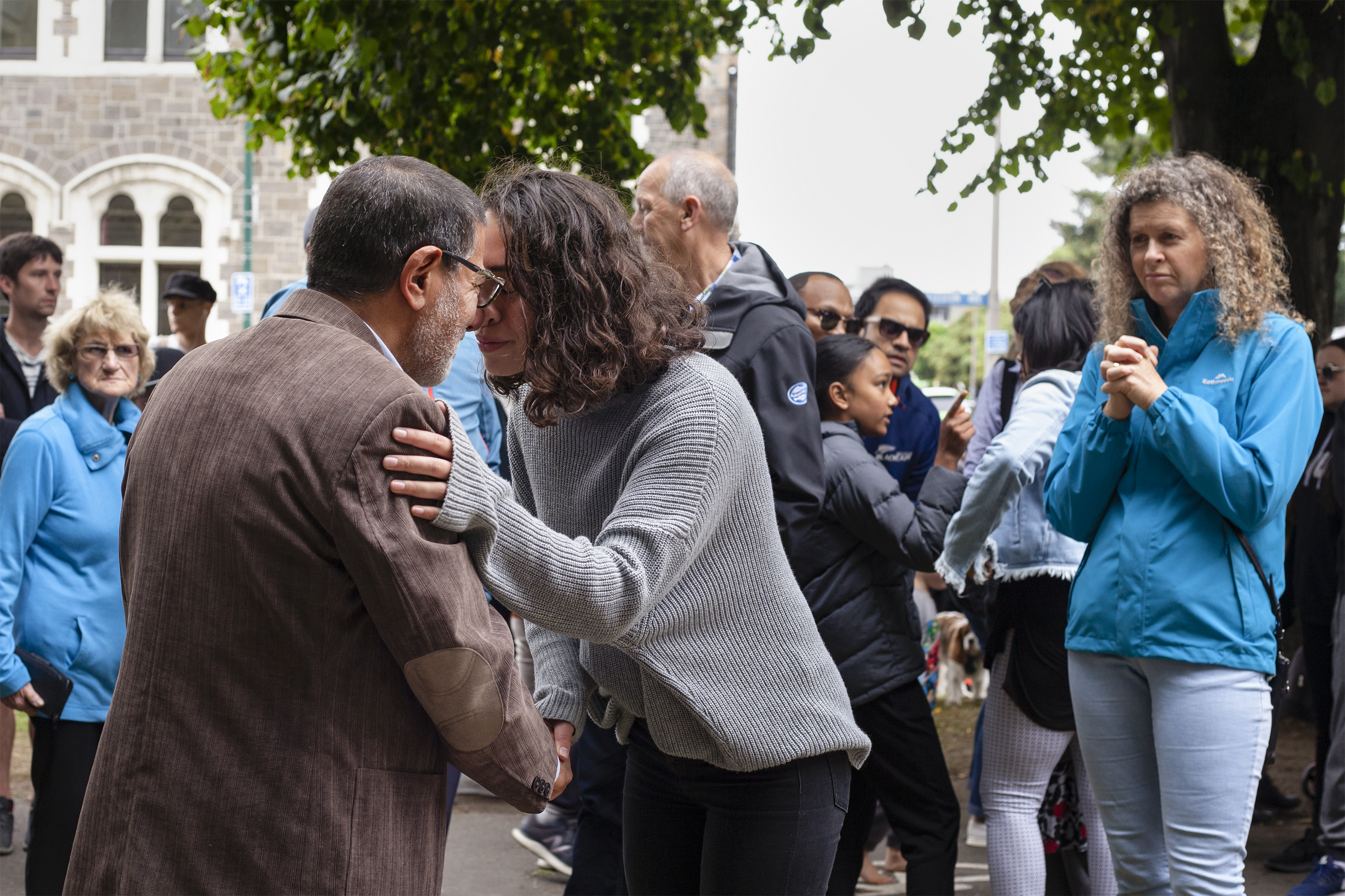 Supporters gather to comfort community members; a woman and man are shown holding hands in a hongi