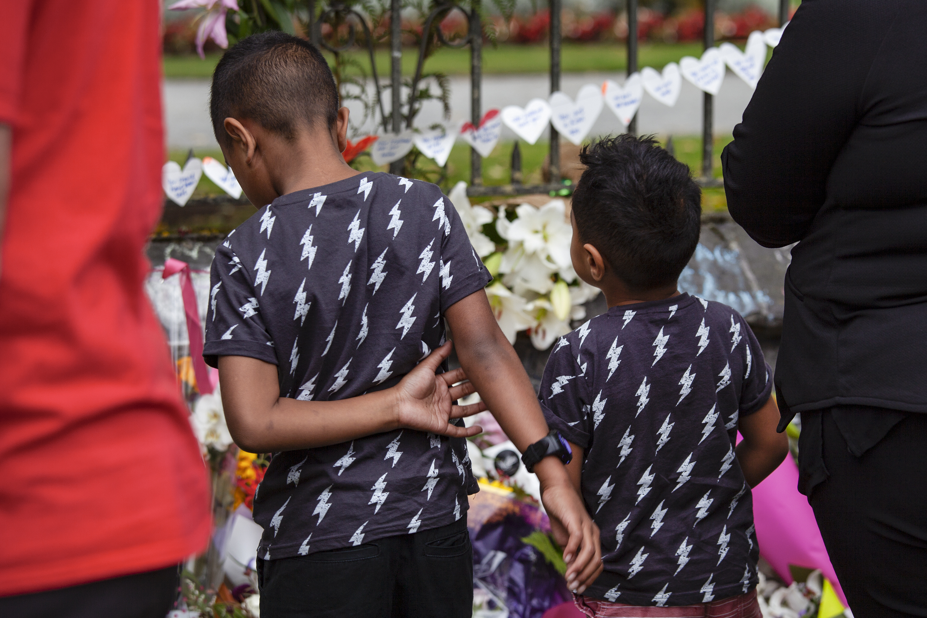 Two young boys in matching lightning bolt shirts stand with adults and view the flowers and messages.
